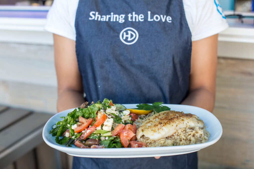 A server wearing a “Sharing the Love” apron from Hunky Dory – an iWS Australia franchise – presents a wholesome plate of grilled fish, brown rice, and a fresh garden salad. The healthy and balanced dish reflects Hunky Dory’s nutritious seafood offerings.