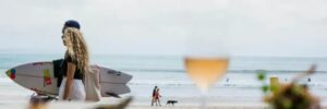 Beach scene with a couple carrying a surfboard, people walking along the shoreline, and a glass of wine in the foreground, representing iWS Australia Hospitality.