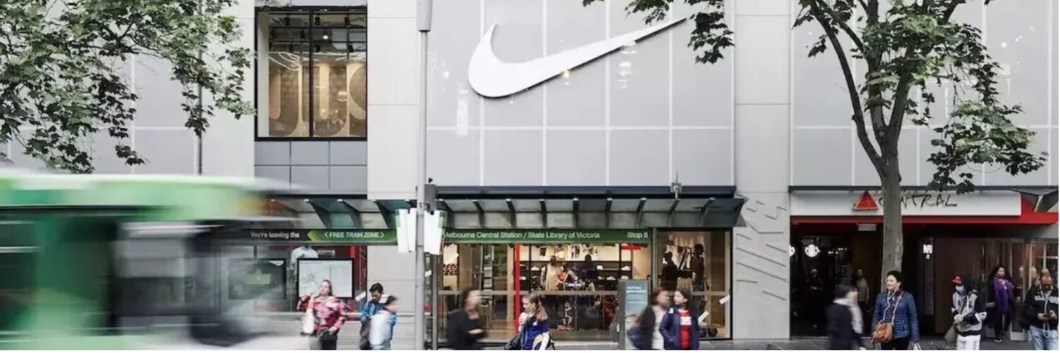 Street view of a Nike retail store with a large white Nike swoosh logo above the entrance and shoppers walking by in an urban shopping area, representing iWS Australia Retailers