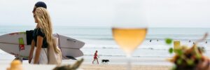 Beach scene with a couple carrying a surfboard, people walking along the shoreline, and a glass of wine in the foreground, representing iWS Australia Hospitality.